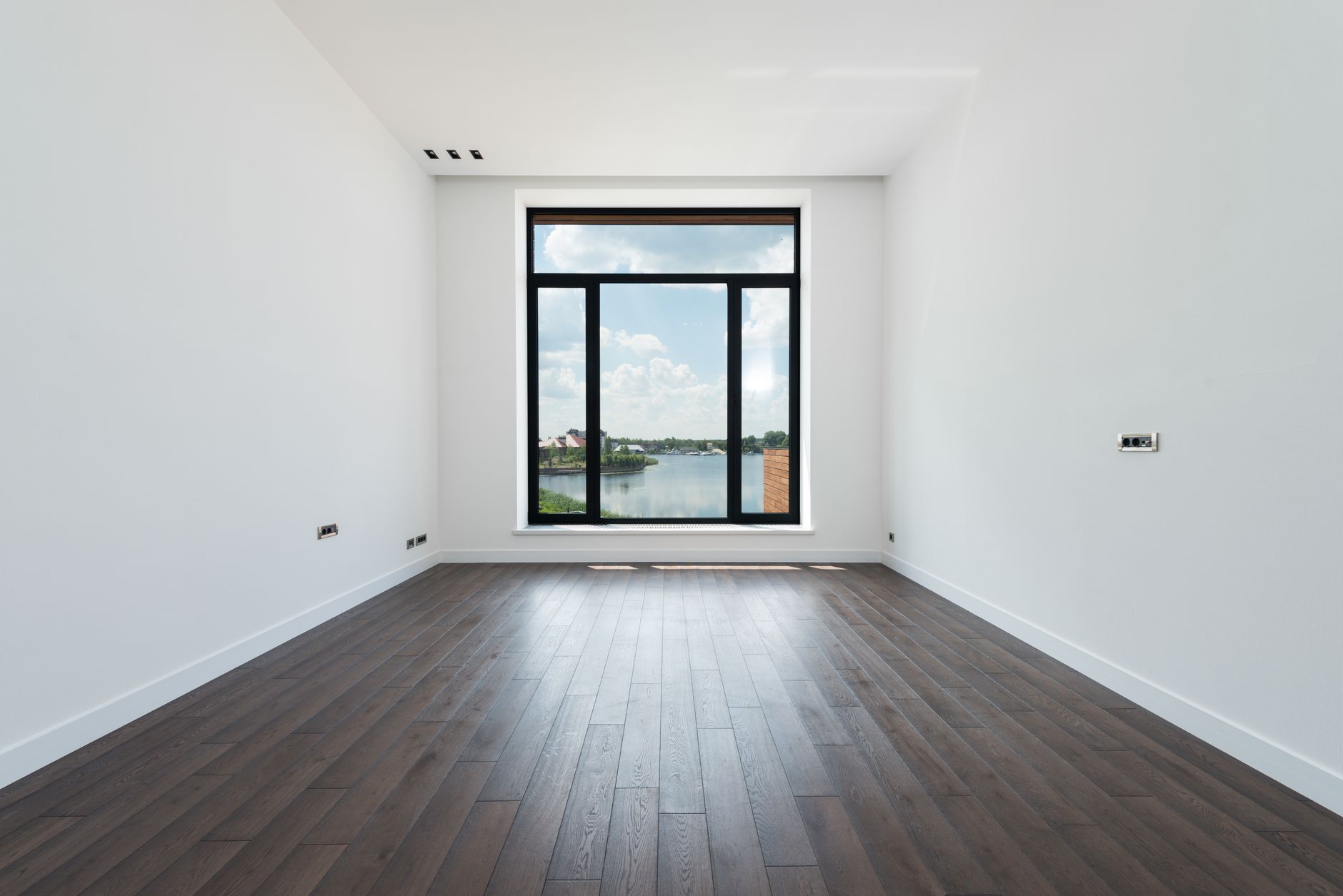 Habitación luminosa con suelo de madera y una gran ventana de marco oscuro que enmarca un paisaje de lago y cielo